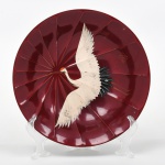 Prato em laca japonesa, formato circular com decoração em relevo radial, fundo em tonalidade vermelha; ornado com representação de ave (grou/tsuru) em voo, executada em técnica pictórica com detalhes em douração; verso em laca escura com inscrição em caracteres japoneses. Medidas: Diam.