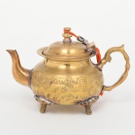 Bule em metal amarelo (latão), com tampa articulada, alça lateral estilizada, bico vertedor curvilíneo, tampa ligeiramente abaulada sob pega torneada. Corpo bojudo com decoração relevada com friso contínuo de motivos geométricos, bico vertedor, pés e alça lateral fixadas por solda. Marcada com inscrição ao fundo. Medidas: