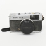 OLYMPUS, modelo Trip 35. Câmera fotográfica para filme 35 mm, corpo em metal com revestimento frontal em material sintético texturizado, acabamento bicolor. Sistema de foco manual por zonas, com objetiva fixa Olympus D.Zuiko 40 mm f/2.8. Obturador central com controle de exposição automático por célula de selênio, dispensando uso de bateria.Visor óptico com indicação de exposição. Ajustes de abertura e distância focal realizados por anel frontal da objetiva. Avanço de filme manual por alavanca superior e rebobinamento por manivela lateral. Fabricação japonesa, década de 19601970. Estado conforme fotografias, apresenta sinais de uso e do tempo. Não foi testado, sem garantias de funcionamento.