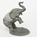 Escultura em bronze fundido, representando elefante em movimento, com tromba elevada e uma das patas anteriores alçadas. Superfície com pátina verde, apresentando variações tonais e marcas de oxidação. Base oval texturizada integrada à composição. Modelagem com tratamento naturalista dos volumes, evidenciando pregas cutâneas, orelhas e detalhes anatômicos. Medidas: