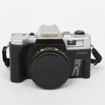 Câmera fotográfica analógica para filme 35 mm, marca YASHICA, fabricação japonesa; corpo em material sintético bicolor, visor óptico integrado, flash embutido com acionamento superior, disparador mecânico, seletor superior de funções e sapata para flash externo. Modelo 2000N, com inscrição Zoom Lens, equipada com lente fixa 50 mm f/6.3, sistema focus free, aro frontal com especificações técnicas e tampa protetora destacável; acompanha alça original. Não foi testada, sem garantias de funcionamento.