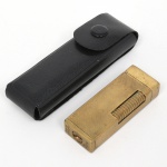 Isqueiro de bolso automático Milo  Automatic De Luxe, em metal dourado, com estojo em material sintético. Corpo retangular de cantos arredondados, estruturado em caixa metálica com tampa articulada superior; faces com decoração em malha quadriculada gravada por estampagem mecânica. Lateral com rolete serrilhado para acionamento do mecanismo de ignição por fricção; extremidade inferior com elemento circular de ajuste. Inscrição gravada na base Milo e Automatic De Luxe. Estado conforme fotografias. Não foi testado, sem garantias de funcionamento.