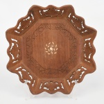 Centro de mesa executado em madeira, de fomato octogonal, com bordo recortado em lobulações contínuas e abas vazadas por recorte fitomorfo, apresentando motivos de folhas estilizadas em repetição. Corpo de perfil baixo e côncavo, com bojo facetado em oito faces inclinadas, convergindo para campo central rebaixado de configuração octogonal. Reserva central ornamentada por marchetaria em composição vegetalista estilizada, circundada por cercadura entalhada em baixo-relevo com motivos florais contínuos. Execução manual, com junções aparentes nos ângulos estruturais. Estado conforme fotografias. Medidas: