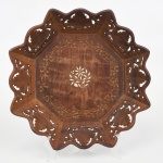 Centro de mesa executado em madeira, de formato octogonal, com bordo recortado em lobulações contínuas e abas vazadas por recorte fitomorfo, apresentando motivos de folhas em composição repetida. Corpo de perfil baixo e côncavo, com bojo facetado em oito faces inclinadas, convergindo para campo central rebaixado de configuração octogonal. Reserva central ornamentada por marchetaria em composição floral estilizada, circundada por cercadura entalhada em baixo-relevo com motivos vegetalistas contínuos. Execução manual, com encaixes visíveis nos ângulos estruturais. Estado conforme fotografias. Medidas: