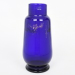 Vaso em vidro soprado de coloração azul cobalto, de corpo cilíndrico alongado com leve inflexão no bojo, assentado sobre base anelar. Ombros suavemente marcados, evoluindo para gargalo cilíndrico curto, com borda lisa e levemente espessada. Superfície com decoração aplicada em douração, em composição floral estilizada disposta em friso no campo superior. Base com marca Esberard Rio. Apresenta desgaste e perdas na douração, e marcas de uso. Medidas: