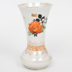 Vaso em porcelana nacional, corpo com bojo inferior elipsoide e gargalo alongado com borda ligeiramente evertida. Decoração por estampagem policromada com motivo floral central em rosa estilizada, predominando as tonalidades laranja, acompanhada por elementos vegetalistas em preto e verde. Faixas ornamentais com douração e padrão linear aplicadas na transição do bojo para o colo e na borda. Marca da manufatura ao fundo. Medidas: