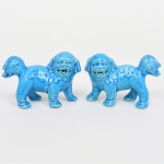 Par de esculturas representando cães de Fó (Shi Shi / leões guardiões imperiais chineses), executadas em porcelana esmaltada. Modelagem em vulto pleno, com corpo compacto apoiado sobre quatro patas, cabeça frontal com mandíbula aberta e dentição aparente; olhos salientes, orelhas pendentes e caudas recurvadas sobre o dorso. Superfície recoberta por esmalte vítreo monocromático em tonalidade azul, com acabamento brilhante. Decoração em relevo com representação estilizada da juba e da pelagem por volutas, espirais e frisos incisos, distribuídos ao longo do dorso, flancos, membros e cauda. Na tradição chinesa, os leões guardiões (Shi Shi) são dispostos em par, associados à proteção simbólica de entradas e espaços, representando equilíbrio complementar. Medidas: Alt.: 5,5 x Comp.: 8,5 x Larg.: 3,5 cm. Apresenta pequeno bicado numa das patas de um deles.