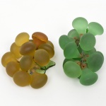 Par de cachos de uva decorativos em vidro fosco, modelados em composição tridimensional, com bagos individualizados em vidro translúcido acetinado nas cores verde e âmbar, fixados por estrutura interna de arame; folhas em vidro moldado com acabamento verde translúcido. Medidas: