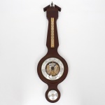 Barômetro de parede com termômetro e higrômetro, caixa em madeira com acabamento envernizado, formato alongado com topo em frontão. Mostrador circular central com mecanismo aneroide visível, escala barométrica graduada; acima, termômetro com escala em graus Celsius; abaixo, higrômetro analógico. Elementos metálicos e mostradores protegidos por visor transparente. Medidas: