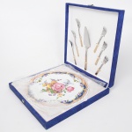 Conjunto para bolo, prato circular em porcelana, aba larga com decoração floral policroma e reservas ornamentais, acompanhado de pá para bolo e seis garfos, cabos metálicos com aplicação decorativa. Sem uso. Medidas: