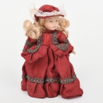 Boneca de coleção em material sintético com indumentária em tecido, com cabeça e membros em material sintético moldado, feições pintadas, olhos fixos e cabelos em fibra sintética de coloração clara, dispostos em cachos. Corpo estruturado, trajando vestido longo em tecido, de modelagem volumétrica, com pregueado na saia, mangas amplas e aplicação de passamanarias. Acompanhada de touca coordenada com aplicação de renda. Estado de conservação conforme fotografias. Medidas: