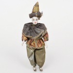 Boneco representando arlequim, com cabeça em porcelana e traje têxtil, com feições estilizadas e policromia aplicada, olhos pintados e lábios definidos em esmalte. Corpo estruturado com enchimento interno e membros em material sintético, mãos e pés moldados. Traje em tecido composto por túnica curta, capa sobreposta e calças amplas, com aplicação de galões decorativos. Indumentária alusiva ao personagem arlequim, com repertório formal vinculado à commedia dellarte. Adereço de cabeça em tecido estruturado, com elemento elevado e aplicação ornamental. Medidas: