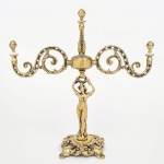Candelabro de três luzes em metal dourado e bronze, com base de formato recortado apresentando elementos em relevo, haste central encimada por elemento torneado e remate superior, sustentado por figura feminina em vulto pleno, da qual partem braços laterais em volutas perfuradas, terminando em suportes para velas(faltando bobeches). Medidas:
