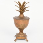 Recipiente com tampa em metal dourado, fundição e conformação, estilo neoclássico com elemento tropical estilizado; corpo em forma de taça com bojo hemisférico, haste curta e base quadrangular com quatro pés em forma de garra, tampa com encaixe ajustado e pega em forma de coroa de folhas dispostas radialmente. Estado de conservação conforme fotografias, com desgaste do acabamento, áreas de oxidação, manchas e marcas de uso. Medidas: