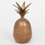 Pote com tampa em metal dourado, com forma decorativa representando abacaxi; corpo ovóide bipartido com encaixe central, superfície com padrão reticulado inciso simulando a casca, interior liso, tampa com encaixe ajustado e pega em forma de coroa de folhas estilizadas dispostas radialmente. Estado conforme fotografias. Medidas: