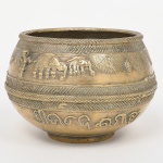 Vaso confeccionado em metal amarelo (latão), manufatura por fundição e cinzelado complementar. Corpo globular com leve contração superior, boca circular com bordo espessado e friso inciso de padrão de corda entrelaçada. Bojo estruturado em faixas horizontais com decoração em relevo e incisão: registro superior com cenas figurativas estilizadas, incluindo figuras antropomórficas, quadrúpedes de corpo alongado e elementos arbóreos; faixa intermediária com motivos geométricos entrecruzados e friso em padrão de espinha; registro inferior com caracteres em relevo de tipologia não latina. Base com pé anelar destacado.Produção de matriz indo-islâmica ou sul-asiática, com repertório iconográfico e ornamental associado à tradição popular regional. Estado conforme fotografias, necessita polimento. Medidas: