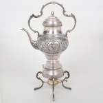 Samovar em metal prateado, composto por reservatório com corpo bojudo decorado em relevo com motivos vegetalistas, bico vertedor curvilíneo, tampa com puxador superior e alça móvel arqueada com ornamentação em relevo, assentado sobre base com compartimento para aquecimento e suporte tripé com pés recortados, apresentando reservatório inferior para queimador. Estado de conservação conforme fotografias, necessita polimento. Medidas: