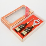 Conjunto composto por saca-rolhas e abridor, em metal e material sintético, com cabos em forma de garrafa, acondicionado em estojo de papelão com visor plástico; saca-rolhas com haste helicoidal metálica e empunhadura modelada, abridor com corpo vazado e elemento de alavanca, ambos com representação de garrafa com rótulo impresso. Medidas: