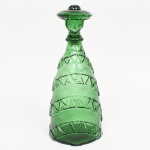 Garrafa de coleção em vidro moldado, de tonalidade verde. Corpo de formato cônico, decorado em faixas horizontais com relevo geométrico em padrão triangular alternado com áreas de textura granulada, formando desenho contínuo que remete a uma espécie de poncho estilizado. Base circular recortada, com marca em relevo MADE IN ITALY. Gargalo curto com anel. Rolha modelada como busto masculino com sombreiro, compondo a figura completa quando encaixada na garrafa. Produção italiana, circa 19501970. Peça de caráter decorativo, com inspiração na indumentária tradicional mexicana. Medidas: Alt.: x Diam.: