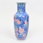 Vaso em porcelana com esmaltação policromada sob vidrado. Corpo ovóide com ombros arredondados, pescoço cilíndrico e borda ligeiramente aberta. Decoração com fundo azul em variação tonal, apresentando motivos florais e ramos com folhas estilizadas, incluindo elementos que remetem a peônias e cachos de frutos. Faixa decorativa na base com padrão repetitivo em vermelho e azul. Interior esmaltado em branco. Provavelmente de fabricação chinesa. Medidas: Alt.: x Diam.: