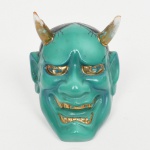 Cabeça de Oni executada em porcelana esmaltada(okimono / ornamento). Escultura em porcelana moldada representando face de oni (demônio do folclore japonês), com expressão marcada, dentes expostos e dois chifres superiores. Modelagem volumétrica com traços acentuados, sobrancelhas arqueadas, olhos amendoados e boca entreaberta com dentes expostos. Superfície com esmaltação em verde-turquesa no rosto, áreas em negro no cabelo e detalhes realçados a douração nos olhos, dentes e base dos chifres. Verso vazado, indicando uso como peça decorativa de parede ou elemento ornamental não funcional (okimono adaptado para fixação/apoio). Medidas: Alt.: 7 x Comp.: 5 x Larg.: 4 cm. Chifres apresenetam pequena quebradura, desgastes na douradura.