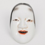 Máscara japonesa em porcelana esmaltada (tipo Noh / Okame), moldada representando rosto feminino estilizado, de feições serenas, olhos semicerrados e boca levemente entreaberta. Cabelos indicados por esmaltação de tonalidade preta, delimitados por traço fino; lábios em tonalidade alaranjada com interior escurecido. Modelagem suave, com volumes contínuos e simplificação formal típica de máscaras teatrais japonesas.Verso fechado, com dois orifícios de suspensão na parte superior, indicando uso como elemento decorativo de parede. Produção por moldagem seriada com acabamento manual na pintura. Tipologia associada a máscaras femininas do teatro Noh ou à figura de Okame/Otafuku, comum em peças decorativas de exportação. Medidas: Alt.: 6,5 x Comp.: 4,5 x Larg.: 4 cm.