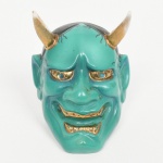 Cabeça de Oni executada em porcelana esmaltada(okimono / ornamento). Escultura em porcelana moldada representando face de oni (demônio do folclore japonês), com expressão marcada, dentes expostos e dois chifres superiores. Modelagem volumétrica com traços acentuados, sobrancelhas arqueadas, olhos amendoados e boca entreaberta com dentes expostos. Superfície com esmaltação em verde-turquesa no rosto, áreas em negro no cabelo e detalhes realçados a douração nos olhos, dentes e base dos chifres. Verso vazado, indicando uso como peça decorativa de parede ou elemento ornamental não funcional (okimono adaptado para fixação/apoio). Medidas: Alt.: 7 x Comp.: 5 x Larg.: 4 cm. Apresenta desgaste na douradura dos chifres.