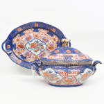 Terrina com tampa e travessa em porcelana esmaltada, China, século XX (segunda metade), estilo de inspiração Imari (revival europeu / exportação)Conjunto composto por terrina coberta e travessa oval com asas vazadas, executado em porcelana de pasta branca, com esmaltação vítrea e decoração policroma sob e sobre esmalte, com acentuado emprego de douração.A terrina apresenta corpo de perfil elipsoidal abaulado, com borda ligeiramente extrovertida e encaixe reentrante para a tampa. Possui duas asas laterais simétricas, moldadas em volutas ascendentes. Tampa de perfil convexo rebaixado, encimada por pegador modelado em forma de botão foliáceo, integralmente dourado.A travessa, de contorno oval lobulado, possui abas laterais com reservas vazadas (pegas integradas), acompanhando o mesmo vocabulário formal da terrina.A decoração organiza-se em campos compartimentados delimitados por cartelas recortadas, contendo composições florais policromas (rosas, crisântemos e flores estilizadas) delineadas a ouro. Os campos são intercalados por fundos densamente pontilhados em vermelho-ferro (aka) sobre esmalte branco, além de extensas áreas em azul cobalto (bleu de four) enriquecidas por salpicos e arabescos dourados (kinrande).O conjunto revela clara filiação ao estilo Imari japonês reinterpretado, com hibridização formal de tradição europeia (tipologia de terrina e travessa de serviço) e vocabulário decorativo orientalizante, típico de produções destinadas à exportação.Interior com esmalte branco levemente esverdeado, característico de porcelana industrial contemporânea.Base da travessa com marca em vermelho sob esmalte: MADE IN CHINA acompanhada de caracteres chineses, indicativa de produção seriada para exportação.Exuberante sopeira executada em porcelana, ao gosto do estilo Imari