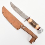 Faca de caça com lâmina em aço, com fio principal liso e dorso parcialmente serrilhado, apresentando marca MUNDIAL gravada. Lâmina de perfil alongado com ponta aguda.Empunhadura em material sintético com aspecto de chifre, com superfície texturizada, intercalada por anéis decorativos nas cores preta e vermelha, terminando em pomo metálico. Guarda metálica reta. Acompanha bainha em couro, com costura aparente, rebites metálicos e passador para cinto, decorada com gravação em relevo. Estado de conservação conforme fotografias. Medidas: