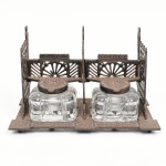 Tinteiro duplo em metal com recipientes em vidro, composto por estrutura em metal fundido, com base retangular e elementos verticais vazados, decorados com motivos geométricos e estilização de leques, formando compartimentos simétricos. Apresenta duas tampas articuladas em metal, de formato circular, destinadas à vedação dos recipientes. Interior com dois reservatórios removíveis em vidro moldado, de seção facetada. Estado conforme fotografias. Medidas: