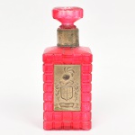 Garrafa em vidro colorido, de corpo prismático, em vidro moldado translúcido na cor vermelha, com superfície estruturada por ressaltos geométricos repetidos. Apresenta gargalo cilíndrico com colar em metal e tampa com pega facetada. Face principal com aplique metálico retangular, em relevo, decorado com brasão heráldico encimado por elmo, ladeado por volutas. Medidas: