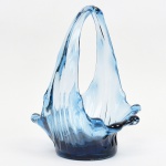 Cesta executada em vidro soprado e moldado, composição orgânica, tonalidae azul translúcido, com modelagem livre, evocando a dinâmica de respingo líquido (efeito splash). Corpo com paredes ondulantes e borda irregular em movimento ascensional. Alça formada por hastes estiradas unidas no ápice. Superfície com estrias do processo de sopro. Medidas: