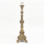 Base de abajur em metal fundido com acabamento dourado. Fuste de composição segmentada, com alternância de elementos torneados e registros decorativos em relevo. Haste central com corpo prismático e aplicações de volutas, cartelas e motivos fitomorfos. Base de configuração trapeziforme com recortes, apoiada sobre três pés em voluta, com decoração em relevo composta por cartelas ovais, frisos e elementos vegetalistas. Colo superior com prato de suporte e soquete em material sintético, associado a instalação elétrica.Estado de conservação com desgaste generalizado do acabamento e sinais de oxidação.