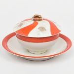Manteigueira em porcelana japonesa, bojo hemisférico fixado sobre presentoir, tampa ligeiramente abaulada encimada por pega superior moldada em dupla folhagem. Esmaltação de tom marfim, dotada de decoração em reservas verticais alternando faixas onduladas em esmalte vermelho-coral e painéis com motivos florais estilizados em douração linear, distribuídos de forma contínua na tampa. Marca em esmalte na base com inscrição MARCA REGISTRADA  KTR  MADE IN JAPAN, indicando produção japonesa de exportação. Medidas: Alt.: 8 x Diam.: 14 cm. Apresenta pequeno bicado na borda do presentoir, e um ínfimo lascado no topo da pega.