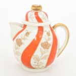 Leiteira em porcelana japonesa, bojo globular assentado sobre base circular,  bico vertedor de seção elíptica e asa lateral em arco. Tampa campaniforme com encaixe interno e pega superior moldada em dupla folhagem. Esmaltação de tom marfim, dotada de decoração em reservas verticais alternando faixas onduladas em esmalte vermelho-coral e painéis com motivos florais estilizados em douração linear, distribuídos de forma contínua pelo corpo e tampa; realçado por generosos arremates de filetes dourados acompanhando bordas, bico e contorno da asa.Marca em esmalte na base com inscrição MARCA REGISTRADA  KTR  MADE IN JAPAN, indicando produção japonesa de exportação. Medidas: Alt.: 13 x Comp.: 12 x Larg.: 9 cm.