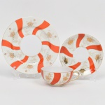 Trio para chá em porcelana japonesa, bojo hemisférico sobre base circular, alça lateral curvada. Esmaltação de tom marfim, dotada de decoração em reservas verticais alternando faixas onduladas em esmalte vermelho-coral e painéis com motivos florais estilizados em douração linear, distribuídos de forma contínua, realçados com filetes em douradura nas bordas. Padrão decorativo replicado no pires e no prato. Marca em esmalte na base com inscrição MARCA REGISTRADA  KTR  MADE IN JAPAN, indicando produção japonesa de exportação. Medidas: Xícara: Alt.: 5 x Comp.: 12 x Larg.: 10 cm; Pires: Diam.: 14 cm; Prato: Diam.: 18 cm.