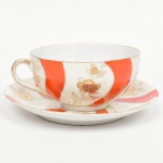 Xícara para chá em porcelana japonesa, bojo hemisférico sobre base circular, alça lateral curvada. Esmaltação de tom marfim, dotada de decoração em reservas verticais alternando faixas onduladas em esmalte vermelho-coral e painéis com motivos florais estilizados em douração linear, distribuídos de forma contínua, realçados com filetes em douradura nas bordas. Padrão decorativo replicado no pires. Marca em esmalte na base com inscrição MARCA REGISTRADA  KTR  MADE IN JAPAN, indicando produção japonesa de exportação. Medidas: Xícara: Alt.: 5 x Comp.: 12 x Larg.: 10 cm; Pires: Diam.: 14 cm.