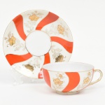 Xícara para chá em porcelana japonesa, bojo hemisférico sobre base circular, alça lateral curvada. Esmaltação de tom marfim, dotada de decoração em reservas verticais alternando faixas onduladas em esmalte vermelho-coral e painéis com motivos florais estilizados em douração linear, distribuídos de forma contínua, realçados com filetes em douradura nas bordas. Padrão decorativo replicado no pires. Marca em esmalte na base com inscrição MARCA REGISTRADA  KTR  MADE IN JAPAN, indicando produção japonesa de exportação. Medidas: Xícara: Alt.: 5 x Comp.: 12 x Larg.: 10 cm; Pires: Diam.: 14 cm.