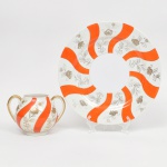 Um(01) açucareiro(falta tampa) e um(01) prato de serviço em porcelana japonesa. Esmaltação de tom marfim, dotada de decoração em reservas verticais alternando faixas onduladas em esmalte vermelho-coral e painéis com motivos florais estilizados em douração linear, distribuídos de forma contínua pelo corpo e tampa; realçado por generosos arremates de filetes dourados acompanhando bordas e contornos dsa asas. Marca em esmalte na base com inscrição "MARCA REGISTRADA - KKR -  MADE IN JAPAN", indicando produção japonesa de exportação. Medidas: Açucareiro: Alt.: 10 x Comp.: 14 x Larg.: 10,5 cm; Prato: Diam.: 25,5 cm.