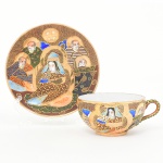 Belíssima xícara para chá em porcelana japonesa com decoração policroma, aplicação de moriage em relevo, realces em ouro e filete na borda. Apresenta composição figurativa formada por uma figura central sentada, acompanhada por quatro Imortais (sennin/rakan), representados como anciãos em trajes tradicionais, tema recorrente em produções de exportação em estilo Satsuma. O fundo do campo decorativo é preenchido por padrão de ondas, com áreas delimitadas por contornos dourados. O verso possui marca em losango contendo a inicial "H" insira em reserva losangular acompanhada de caracteres, identificação utilizada na porcelana japonesa de exportação a partir do início do século XX. A iconografia e a técnica de moriage situam a peça dentro da produção japonesa voltada ao mercado externo entre o final do período Taisho e o início do período Showa. Medidas