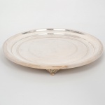 Grande salva confeccionada em metal espessurado à prata, formato circular de centro ligeiramente rebaixado, lavrado com faixas em em guilloché distribuídas ao centro e na borda, assente sobre três pés cinzelados com volutas e folhagens. Medidas: Alt.: 4 x Diam.: 39,5 cm.