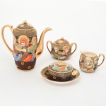 Conjunto executado em cerâmica japonesa com decoração policroma, aplicação de moriage em relevo, realces em ouro e filete nas bordas. Apresenta composição figurativa formada por uma figura central sentada, acompanhada por quatro Imortais (sennin/rakan), representados como anciãos em trajes tradicionais, tema recorrente em produções de exportação em estilo Satsuma. O fundo do campo decorativo é preenchido por padrão de ondas, com áreas delimitadas por contornos dourados. O verso possui marca em losango contendo a inicial "JH" insira em reserva losangular acompanhada da inscrição "made in japan". A iconografia e a técnica de moriage situam a peça dentro da produção japonesa voltada ao mercado externo entre o final do período Taisho e o início do período Showa. Composto de um(01) bule para café, um(01) açucareiro, uma(01) manteigueira e uma(01) cremeira. Medidas: Alt.: 18 x Comp.: 17,5 x Larg.: 8 cm; Açucareiro: Alt.: 9 x Comp.: 13 x Larg.: 8 cm; Manteigueira: Alt.: 7 x Diam.: 13 cm; Cremeira: Alt.: 7,5 x Comp.: 9 x Larg.: 7,5 cm. Açucareiro possui manchas do tempo na parte interna, e craquelado intenso, borda interna de encaixe da tampa com pequeno trincado.
