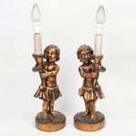 Par de luminárias em madeira entalhada, com acabamento em douração (folheação/pátina dourada), representando figuras infantis em vulto pleno, de caráter putti, derivadas da tradição decorativa europeia. As figuras apresentam modelagem volumétrica, cabelos ondulados em relevo e vestem saia estilizada, apoiando-se sobre base circular com superfície texturizada e motivos vegetalistas em relevo. Cada figura sustenta haste vertical integrada, encimada por suporte para lâmpada em formato de vela falsa, eletrificada.Bases circulares com topo trabalhado em relevo simulando terreno orgânico; hastes integradas ao corpo escultórico; soquetes superiores adaptados para instalação elétrica. Parte elétrica funcionando. Medidas: Alt.: 59 x Diam.: 15 cm.