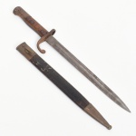Baioneta militar com bainha, manufatura Weyersberg Kirschbaum & Cie., Solingen, Alemanha((fundada em 1584 em Solingen), final do século XIX - início do século XX; lâmina reta confeccionada em aço, dotada de canal longitudinal central (gola), montada com guarda simples em ferro provida de gancho curvo. Empunhadura composta por talas de madeira fixadas por rebites metálicos sobre espiga de aço, conserva sua bainha original, constituída por corpo em metal com ponteira e bocal metálicos; base da lâmina encontra-se estampada a marca WEYERSBERG KIRSCHBAUM & CIE  SOLINGEN, tradicional fabricante alemão estabelecido na cidade de Solingen, importante centro europeu de produção de lâminas militares desde o século XIX. A empresa forneceu espadas, sabres e baionetas para diversos exércitos europeus e latino-americanos. Peça de tipologia militar regulamentar, destinada originalmente ao acoplamento em fuzil por meio do sistema de encaixe localizado no pomo do punho. Estado conforme fotografias. Medida: Comp.: 45 cm.
