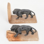 Par de serre livre composto por esculturas de leões em metal fundido, apoiadas sobre bases retangulares em alabastro. Figuras representadas em marcha, com juba estilizada e cauda arqueada. Bases com elemento vertical de contenção em uma das peças. Apresentam desgastes, escoriações e perdas nas bases em alabastro, com lascas e sinais de uso e restauro aparente. Medidas: Alt.: 15 x Comp.: 22 x Larg.: 9 cm. Alt.: 10,5 x Comp.: 22 x Larg.: 9 cm.