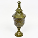 Pote com tampa em cerâmica moldada, de corpo ovóide com leve constrição no colo, assente sobre pé anelar destacado; bordo espessado com assento interno para encaixe da tampa; tampa ajustada, de perfil campanular, encimada por pega em botão; decoração em relevo aplicado com motivos fitomorfos em guirlandas e frisos concêntricos; vidrado monocromático em tonalidade verde oliva, de acabamento vítreo, com variações de queima. Medidas: