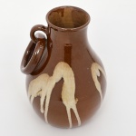 Jarro em cerâmica vidrada, com bojo globular, colo estreito e bordo simples ligeiramente evertido. Apresenta alça lateral aplicada de seção circular, configurada em anel. Superfície recoberta por vidrado marrom homogêneo, com decoração em escorridos (dripping) em tom bege, resultantes de aplicação de engobe/vidrado contrastante sob efeito de gravidade durante a queima. Base plana não vidrada. Medidas: