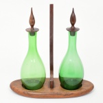 Galheteiro composto por duas galhetas em vidro verde, de corpo bojudo e gargalo alongado, com tampas em madeira torneada, acondicionado em suporte de madeira com base oval e divisória central vertical. Medidas: