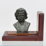 Um(01) serre livre em madeira com busto em pétit bronze  representando o compositor brasileiro Carlos Gomes(1836 -.1896). Base retangular em madeira maciça com elemento vertical de contenção; sobre esta, placa superior sustentando o busto moldado, com identificação em relevo na base frontal. Medidas: Alt.: 16x Comp.: 19 x Larg.: 11 cm.