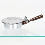 Cata-migalhas manual em metal prateado, formato circular com compartimento coletor e tampa articulada; superfície externa com brasão em relevo de caráter decorativo. Cabo em madeira torneada, fixado por suporte metálico, permitindo o acionamento manual do mecanismo de recolhimento. Utensílio de mesa destinado à remoção de migalhas. Apresenta sinais de oxidação.